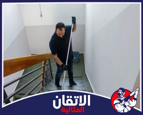 تنظيف مداخل المباني التجارية بالقطيف 0567008659 الأتقان المثالية عامل يقوم بتنظيف درجات مدخل عمارة تجارية في القطيف باستخدام ممسحة طويلة لضمان نظافة الأرضيات وسلامة الممرات.