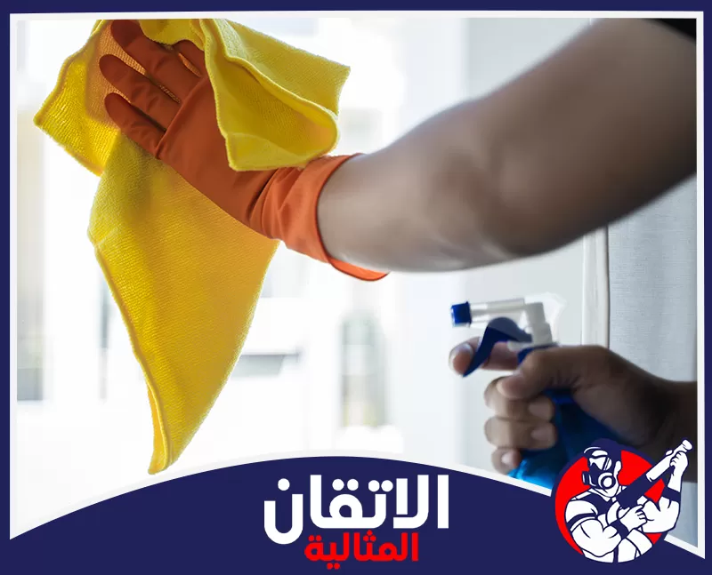تنظيف الدرايش في الدمام 0567008659 شركة أتقان المثالية تُظهر الصورة لقطة مقربة ليد ترتدي قفازًا برتقاليًا وهي تمسح سطحًا زجاجيًا بقطعة قماش صفراء ناعمة.