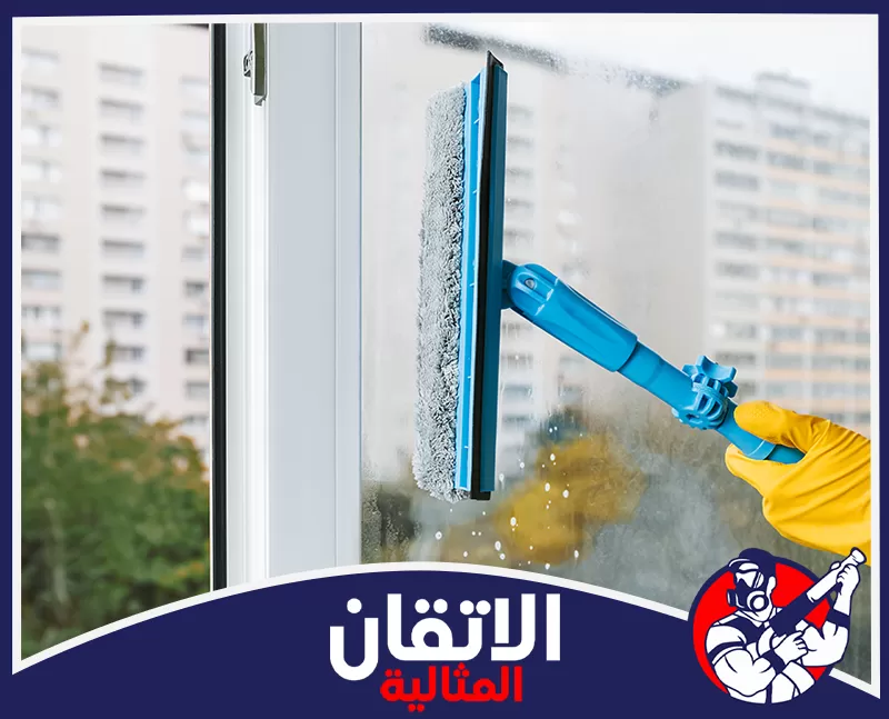 تنظيف الدرايش في الخبر 0567008659 شركة أتقان المثالية يد بقفاز أصفر تنظف نافذة زجاجية بممسحة زرقاء، وتظهر مبانٍ سكنية في الخلفية