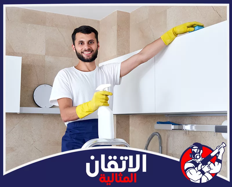 شركة تنظيف مطابخ مجربة بالقطيف 0567008659 شركة أتقان المثالية رجل يرتدي قفازات صفراء، يمسح خزائن المطبخ البيضاء بحماس، ويحمل عبوة منظف وبخاخًا.