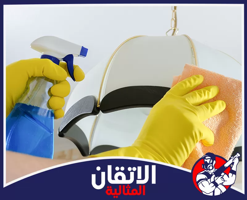 صيانة النجف بالأحساء 0567008659 شركة أتقان المثالية شخص يرتدي قفازات صفراء في وينظف مصباحاً أبيض باستخدام بخاخ وقماش برتقالي.